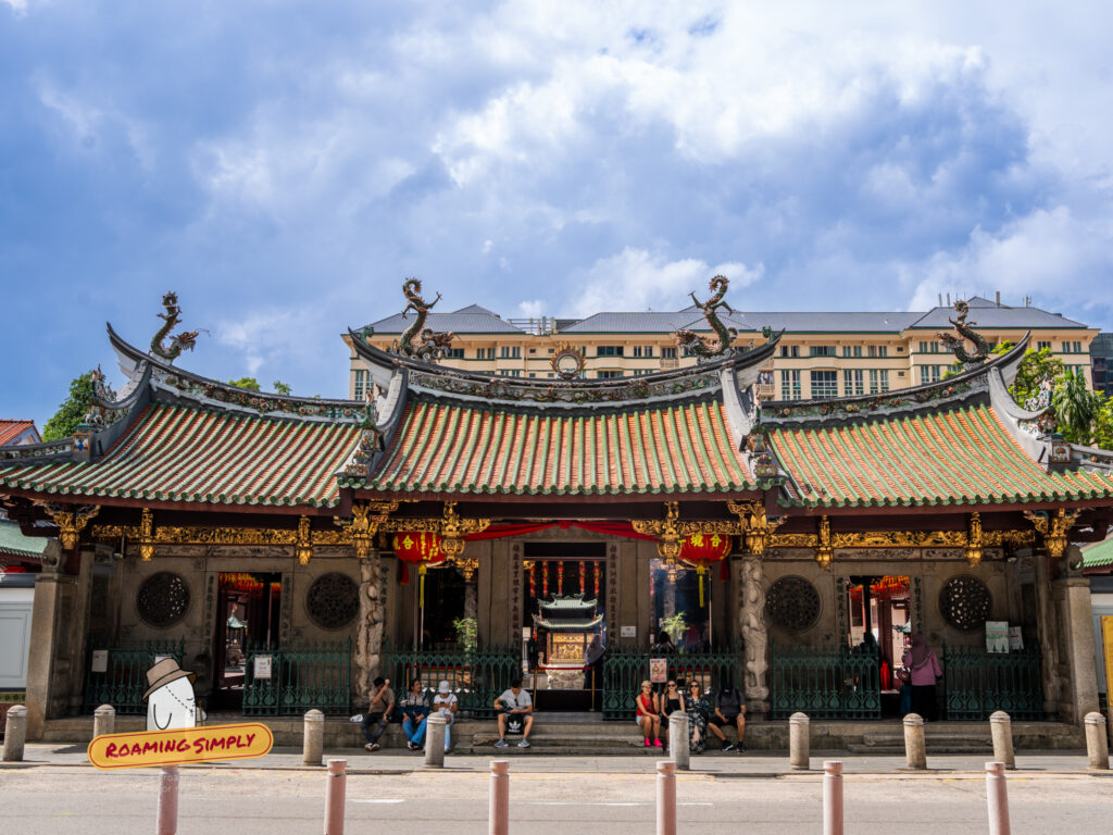 Front facade of Thian Hock Keng Temple in Singapore under a blue sky, featuring intricate green and orange tiled roofs with porcelain dragon sculptures. Visitors are sitting on the stone steps of the entrance, and the traditional architecture contrasts with the modern buildings in the background.