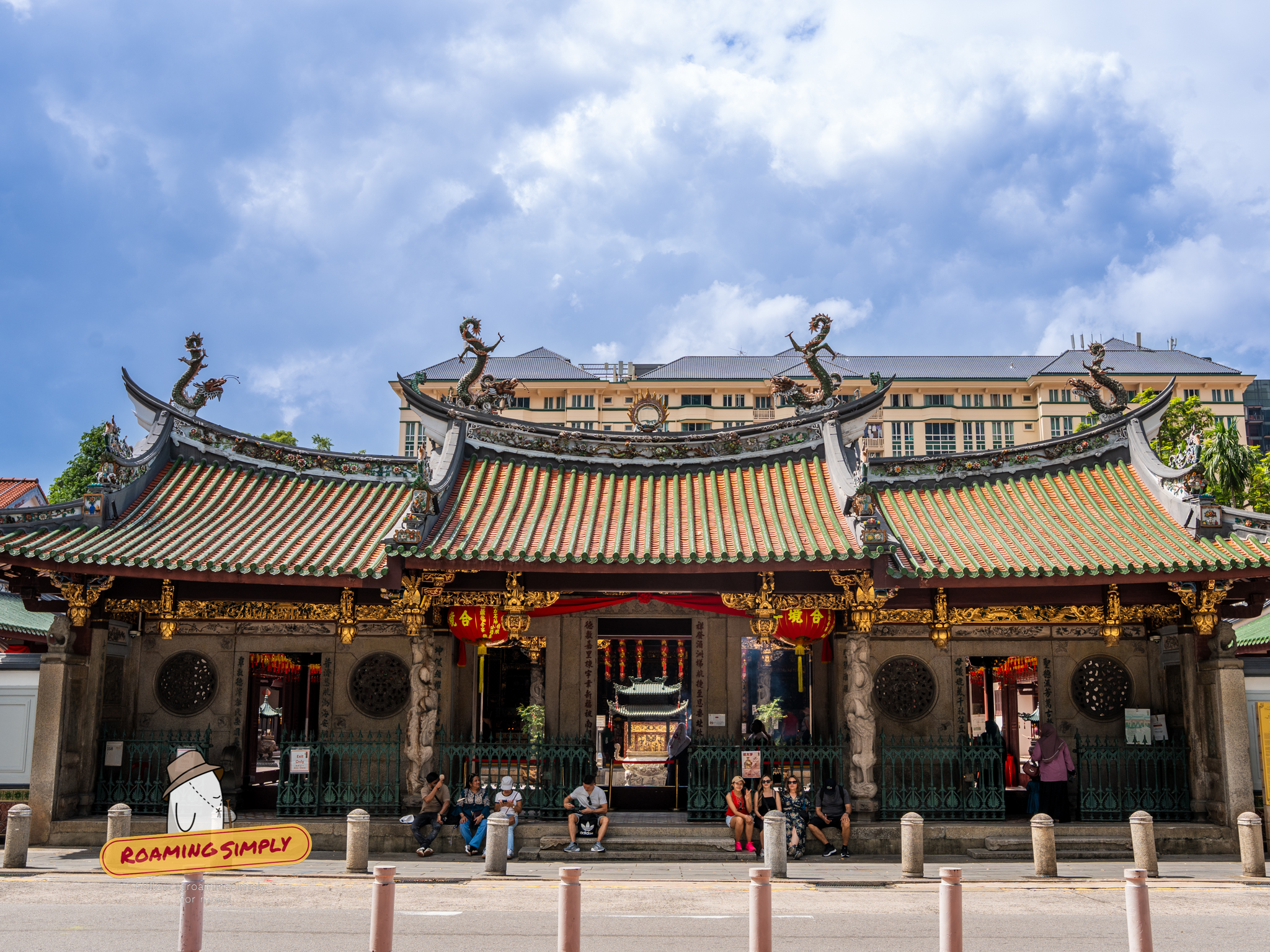 Front facade of Thian Hock Keng Temple in Singapore under a blue sky, featuring intricate green and orange tiled roofs with porcelain dragon sculptures. Visitors are sitting on the stone steps of the entrance, and the traditional architecture contrasts with the modern buildings in the background.