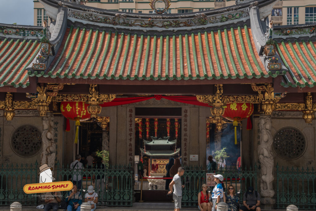 Front entrance of Thian Hock Keng Temple showing the ornate tiled roof, gold-gilded wood carvings, and red lanterns, with visitors gathering at the gate and sitting on the steps.