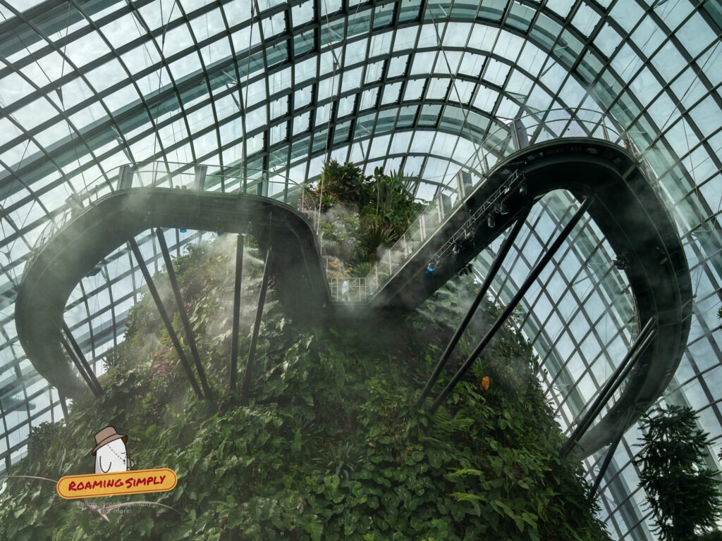 Low-angle view inside the misty Cloud Forest dome at Gardens by the Bay, Singapore, featuring an elevated curved walkway wrapping around a towering indoor mountain covered in lush green plants under a glass ceiling.