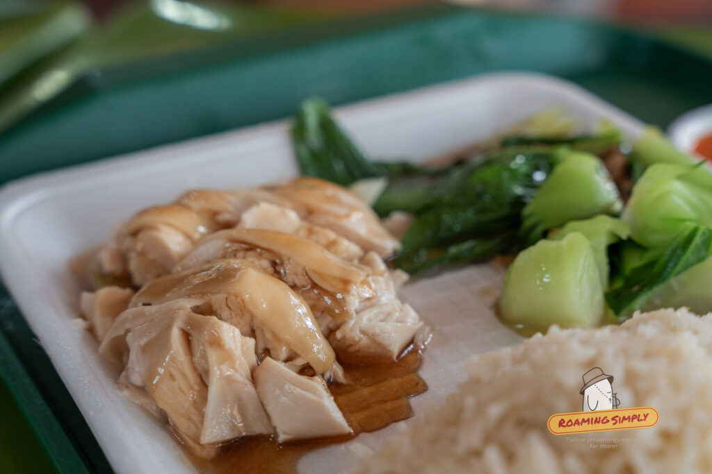 Side angle close-up of poached chicken slices at Ah Tai Chicken Rice, showing the glistening, tender meat and gelatinous skin drizzled with soy sauce, situated next to blanched bok choy.