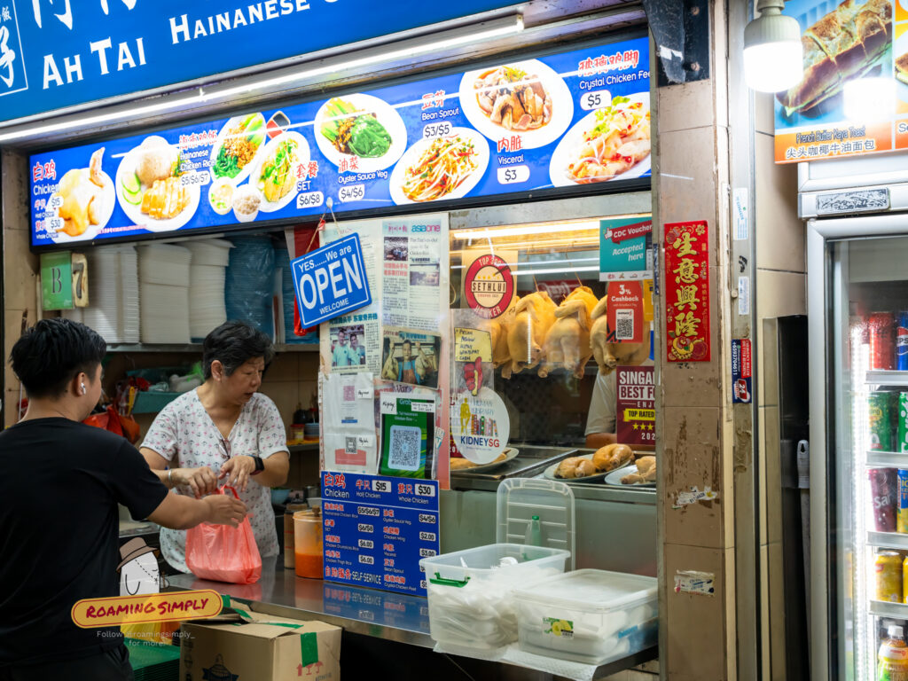 Storefront of Ah Tai Hainanese Chicken Rice at Maxwell Food Centre, featuring a blue signboard with menu photos, hanging poached chickens in the glass display, and a queue of customers.