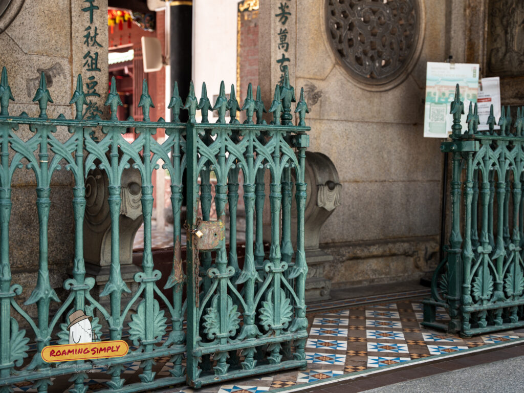 Close-up of a weathered green iron gate with a rusty lock at Thian Hock Keng Temple, set against stone pillars with Chinese calligraphy and vintage geometric floor tiles.