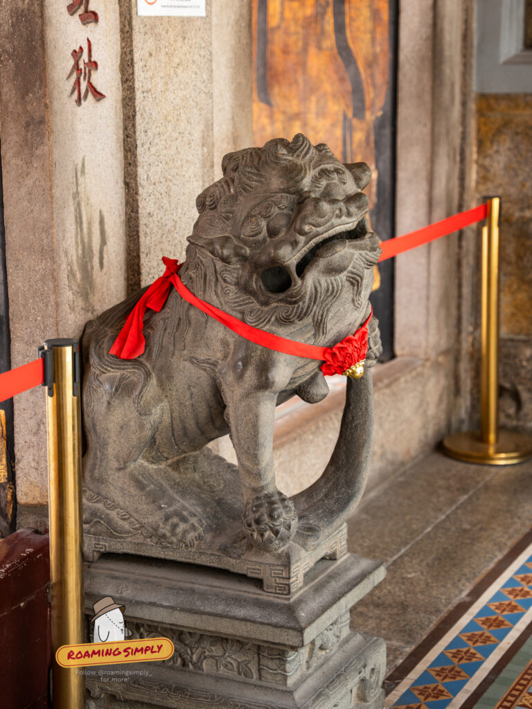 Stone guardian lion statue at the entrance of Thian Hock Keng Temple, adorned with a bright red ribbon sash and sitting on a carved pedestal.