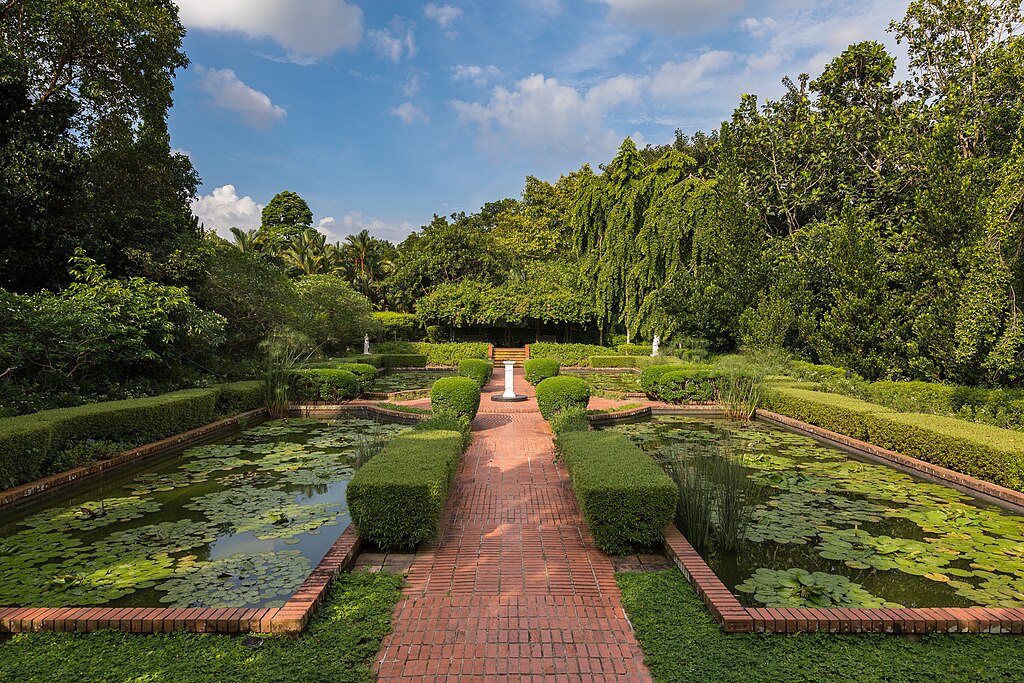Symmetrical view of the formal Sundial Garden in the Singapore Botanic Gardens, featuring a central red brick path flanked by rectangular lily ponds and lush tropical trees.