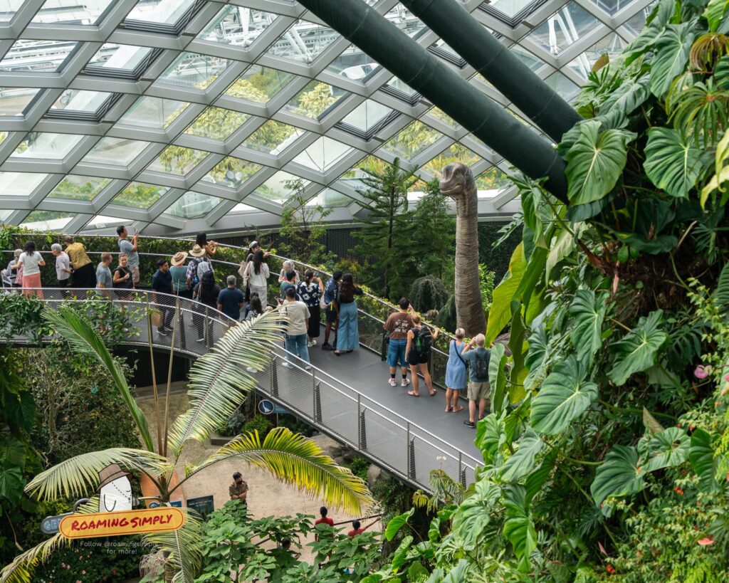 Visitors walking along an elevated metal walkway inside the glass Cloud Forest dome at Gardens by the Bay in Singapore, with a tall dinosaur statue visible among the lush greenery.