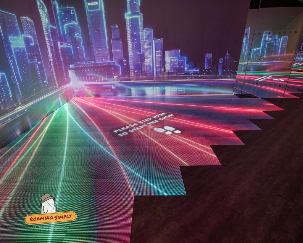 Photograph of an immersive 3D interactive display room at the Singapore City Gallery, featuring neon lights projecting a futuristic city skyline and floor footprints that read 'Please step here to start the show'.