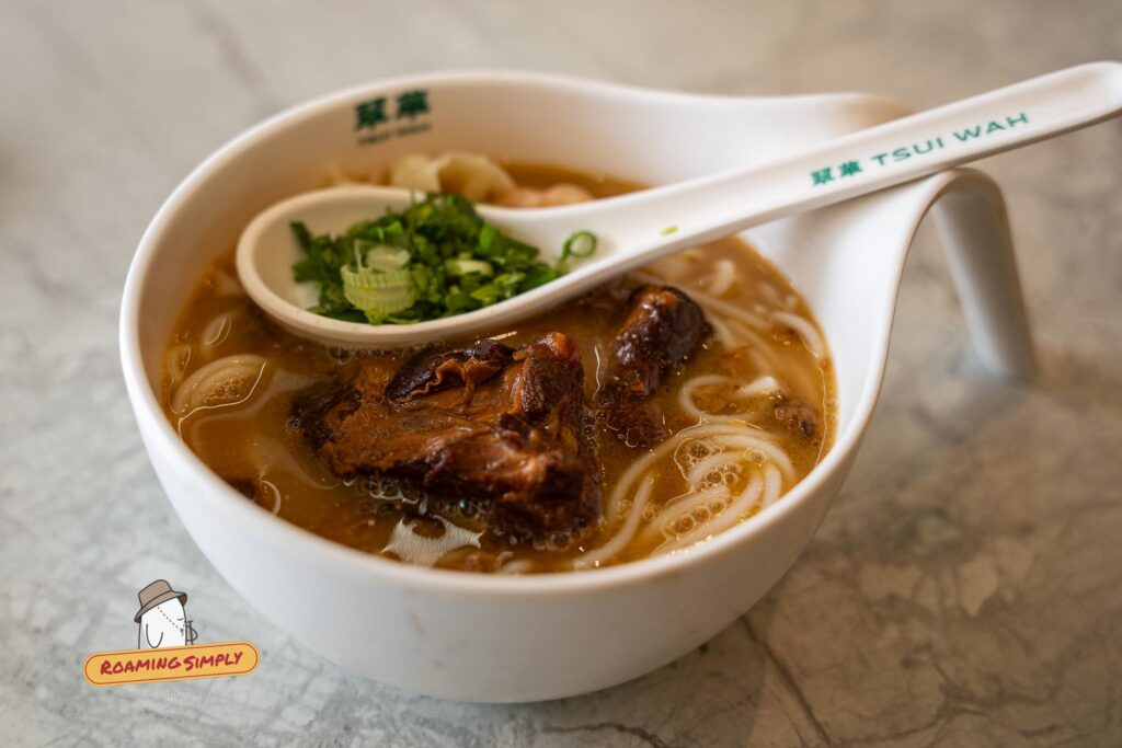Close-up photograph of a white branded Tsui Wah bowl filled with Kagoshima-style pork cartilage noodles in a dark broth, with a matching ceramic spoon holding freshly chopped green onions resting on top.