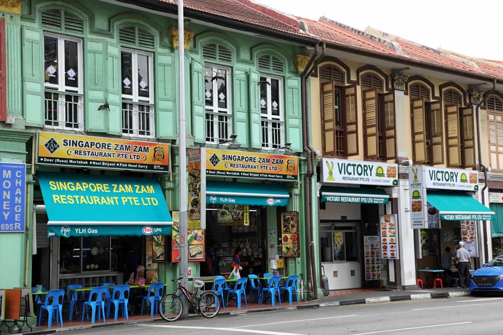 Exterior street-level photograph of the historic Singapore Zam Zam restaurant, featuring a vibrant green traditional shophouse facade, bright blue awnings, and outdoor seating in Kampong Glam.
