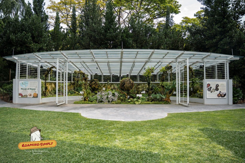 The Hoya House at Singapore Botanic Gardens, a curved open-sided glasshouse with a white steel frame, planted with hoyas and fronted by interpretive signboards on a lawn.