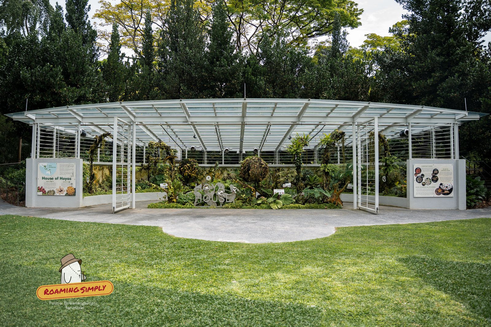 The Hoya House at Singapore Botanic Gardens, a curved open-sided glasshouse with a white steel frame, planted with hoyas and fronted by interpretive signboards on a lawn.