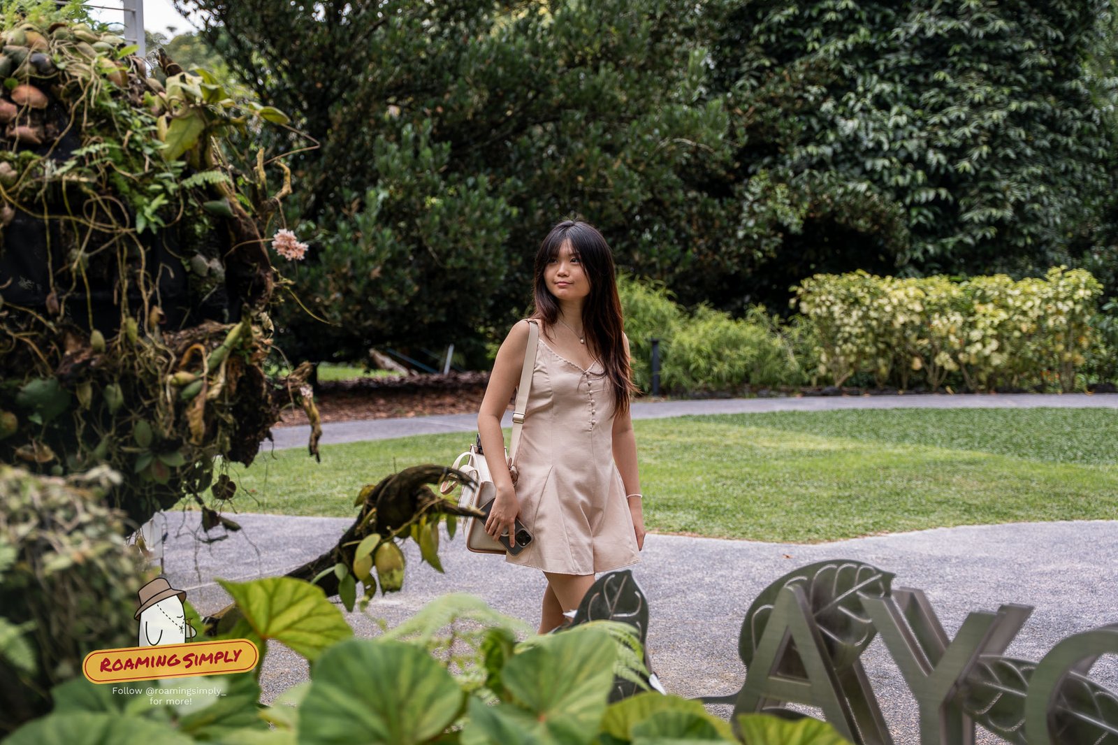 A visitor standing on the path at the Hoya House, Singapore Botanic Gardens, framed by hanging hoya vines and broad green leaves in the foreground.