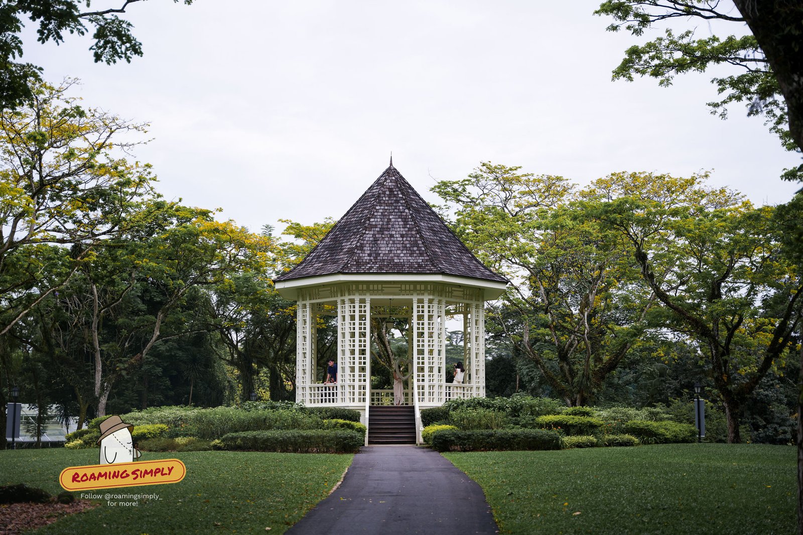 The white octagonal Bandstand at Singapore Botanic Gardens, with its dark shingled roof and lattice pillars, framed by trees at the end of a path across the lawn.