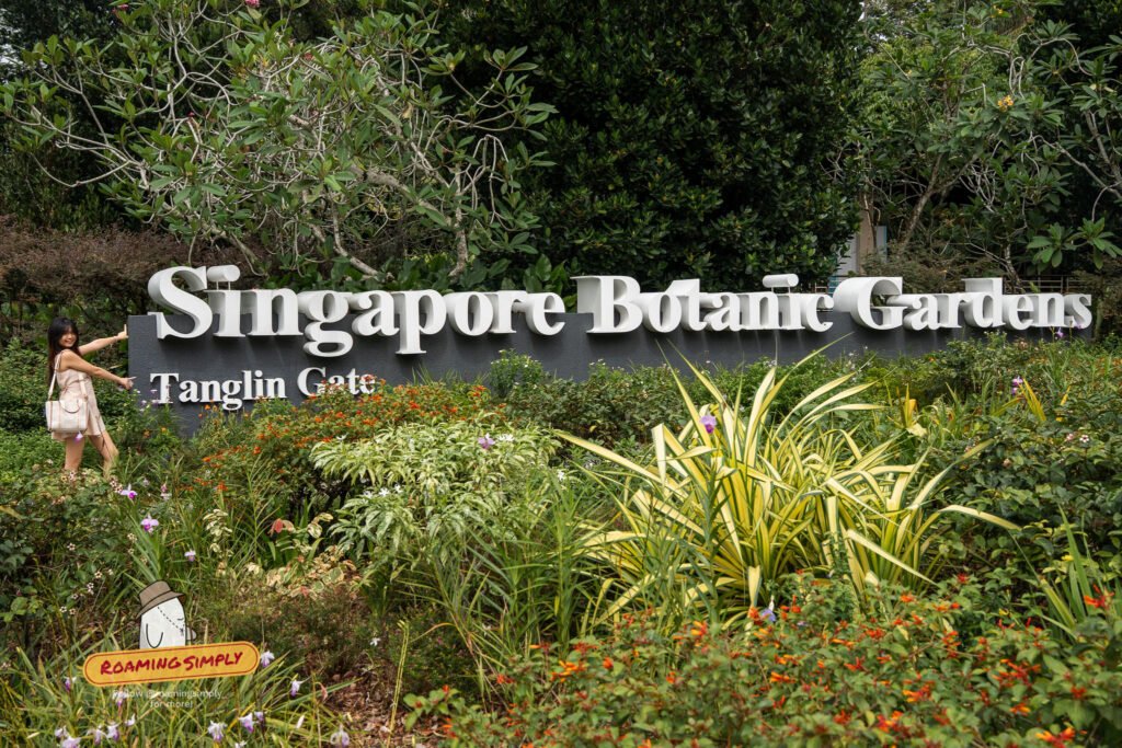 The Tanglin Gate entrance sign at Singapore Botanic Gardens, with a visitor standing beside it among flowering shrubs.