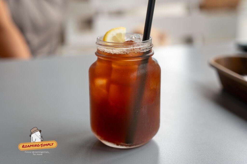 An iced amber drink in a glass jar with a lemon slice and a black straw on a grey table