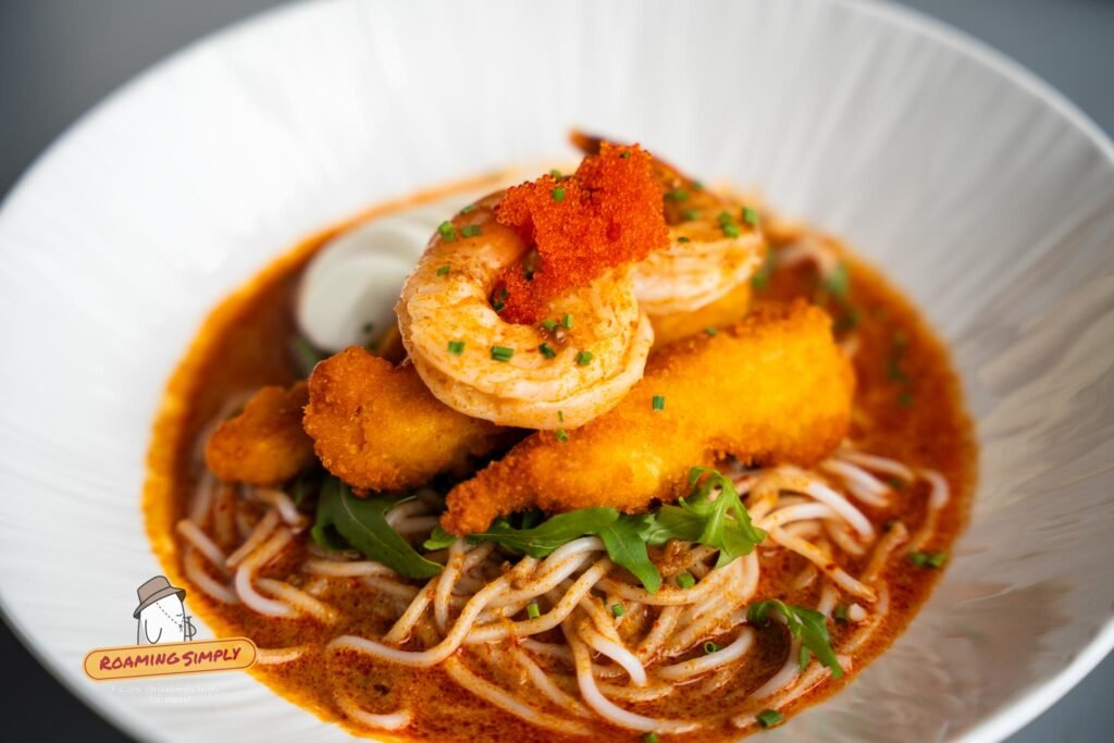 A bowl of laksa with thick rice noodles, prawns, breaded fried pieces, tobiko and rocket leaves