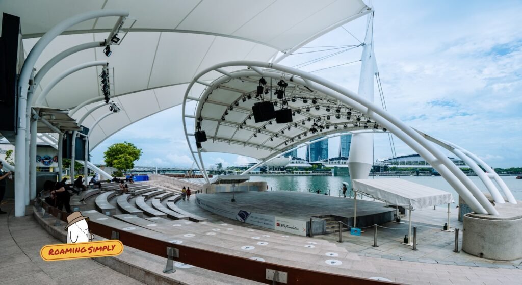 Open-air stage under a white canopy with curved stone seating, facing Marina Bay