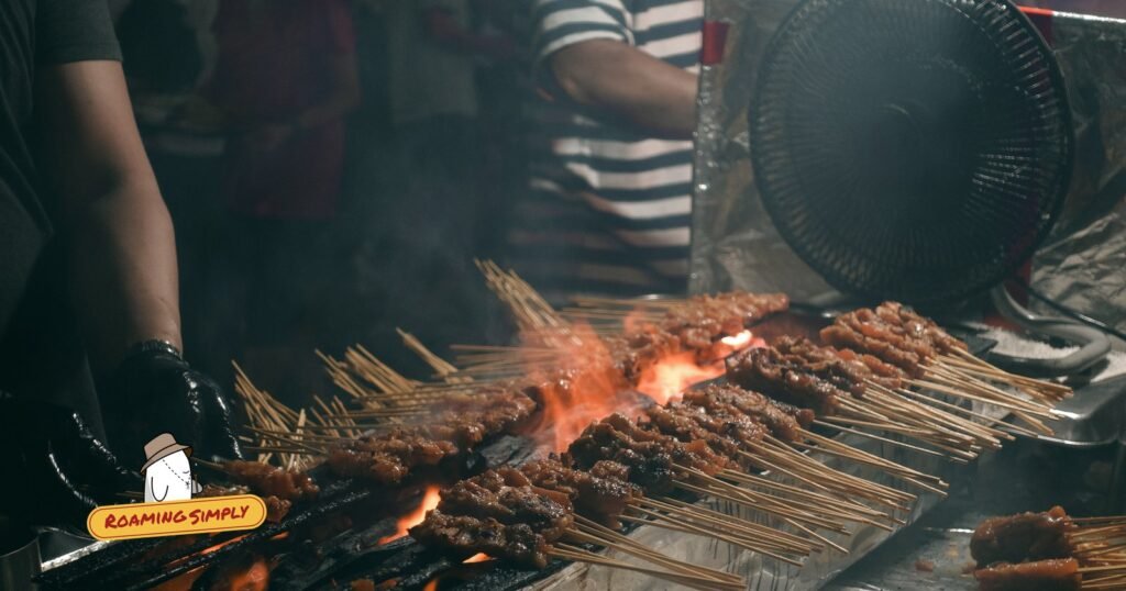 Rows of satay skewers grilling over flames at night, with a gloved hand turning them
