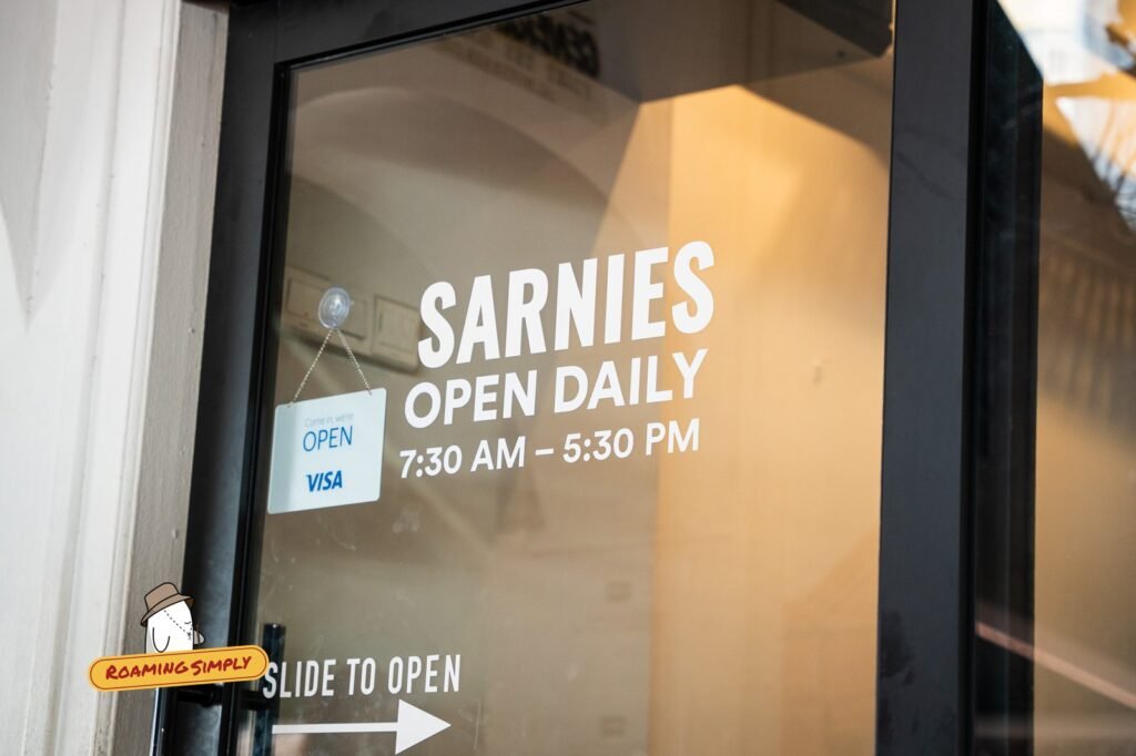 Glass door with white lettering that reads Sarnies, open daily, 7:30 AM to 5:30 PM